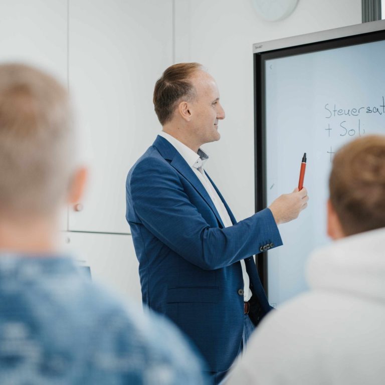 main-lohn - Immer auf dem Laufenden, News Steuerberater steht vor einer digitalen Tafel in den Kanzleiräumen in Großwallstadt und erklärt einer Gruppe von Schülern Lohnabrechnung.